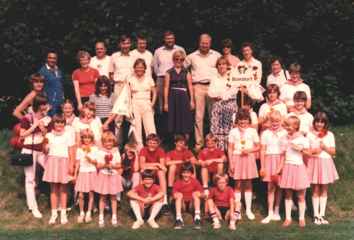 Inhaltsbeschreibung: Gruppenfoto mit 22 Kindern beim Kinderturnfest in Gerlingen 1982. Die Mädchen waren mit weißen T-Shirts und rot-weiß karierten Röckchen gekleidet, die Jungs hatte schwarze Turnhosen und rote T-Shirts an. Mit auf dem Foto Betreuer und mitgereiste Eltern.